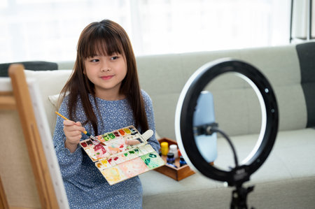 A pretty and happy young Asian girl is live streaming on her social media while painting in the...