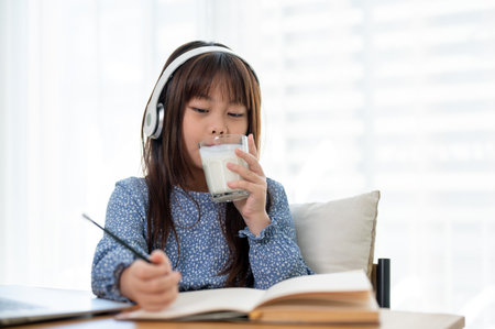 An adorable young Asian girl is drinking a glass of milk while doing homework at a table in her...