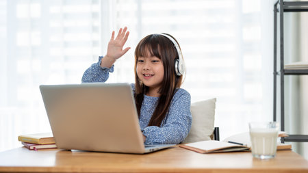 A cheerful and cute young Asian girl is raising her hand to answer a question while studying online...