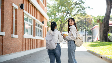 A beautiful and positive young Asian female college student in casual clothes and backpack walking with her friend on a footpath in a campus. Education conceptの写真素材