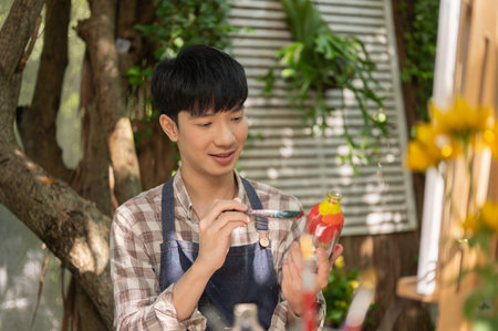 A happy and handsome young Asian man in an apron is coloring a glass bottle, handcrafting a decorative bottle at home. creativity hobby, home leisureの写真素材