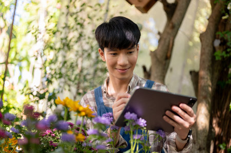 A portrait of a positive handsome Asian male florist or flower gardener is using his digital tablet while working at flower plant nursery in a garden.の写真素材