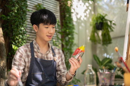 A happy and handsome young Asian man in an apron is coloring a glass bottle, handcrafting a decorative bottle at home. creativity hobby, home leisureの写真素材