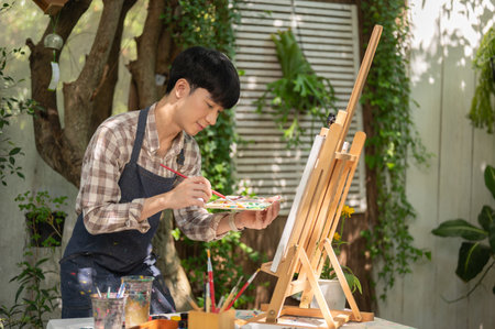 Young happy Asian man artist in an apron is coloring on a canvas on an easel in his home garden. artwork, art workshop, creative activityの写真素材