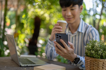 A positive, handsome Asian man is having a coffee and using his smartphone while sitting at a table in a beautiful green garden. People and technology conceptsの写真素材