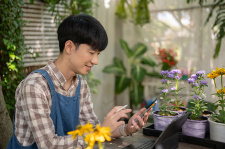 Young positive and handsome Asian male florist or flower shop owner is checking online orders on his smartphone, working in his small garden.の写真素材