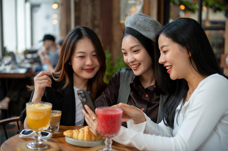 Group of cheerful and attractive Asian female friends are enjoying hanging out at a cafe or restaurant in the city together, chit chat, watching something interesting on a phone together.の写真素材