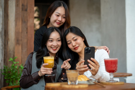 Group of positive and beautiful Asian female friends are enjoying chit chat, gossiping, looking online contents on a smartphone while hanging out at a cafe in the city together.の写真素材