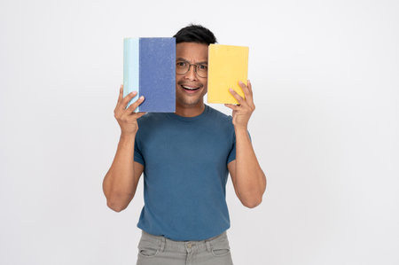 A clueless, doubtful Asian man in casual clothes is posing with books while standing on an isolated...