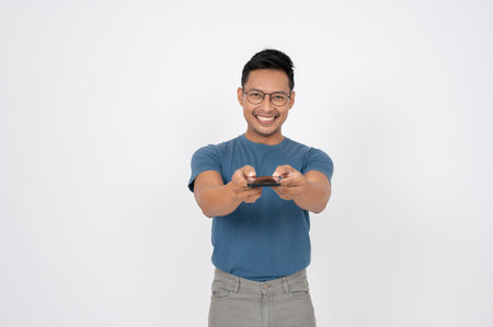 A positive, handsome Asian man in casual clothes is handing, giving his smartphone at the camera, standing on an isolated white background. mobile application, social media, wireless deviceの写真素材