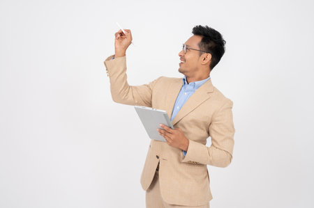 A happy and professional Asian businessman in a formal suit is looking aside at an empty space, standing with his digital tablet on an isolated white background.の写真素材