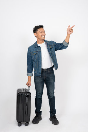 A positive, handsome Asian man in jeans jacket with his luggage is pointing his finger at an empty space on an isolated white background. giving direction, advice, presentationの写真素材