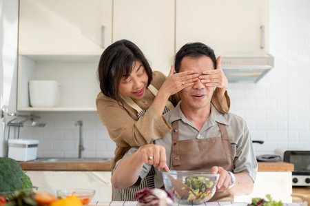 A happy, playful Asian wife is teasing her husband while he is cooking. Peek a boo, covering his eyes, surprise! family bonding, wife and husband, domestic lifeの写真素材