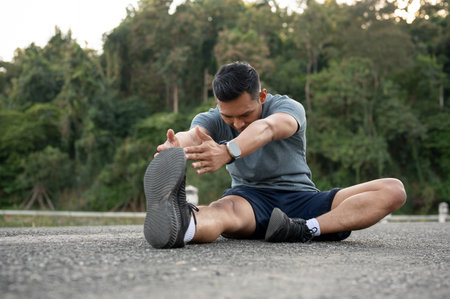 A happy, strong Asian man in sportswear is stretching his body on the floor outdoors, warming up...