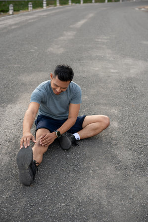 A happy, strong Asian man in sportswear is stretching his body on the floor outdoors, warming up before exercise or cooling down after exercise. runner, jogger, athleteの写真素材