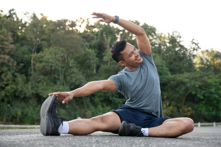 A happy, strong Asian man in sportswear is stretching his body on the floor outdoors, warming up...