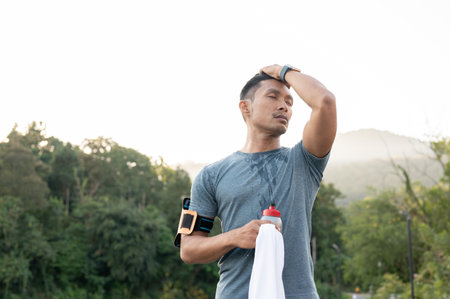 A tired Asian man is wiping his sweat on his face, resting after a long run outdoors in the morning. intense workout conceptの写真素材