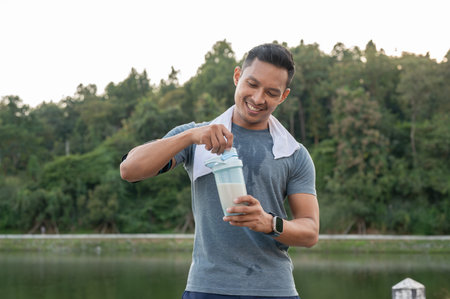 A happy, strong and fit Asian man in sportswear is drinking a protein shake after a long run outdoors, having a sport drink supplement for muscle gain.の写真素材