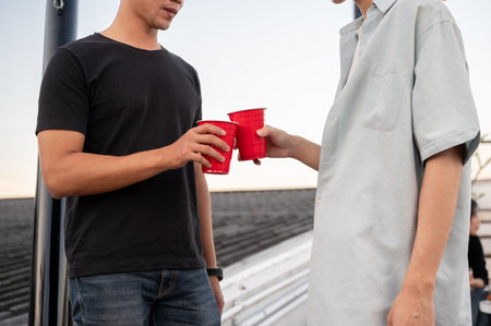 Two interesting young Asian male friends are toasting beers and enjoying talking while partying on a rooftop bar together. Friendship and night life concepts. close-up imageの写真素材