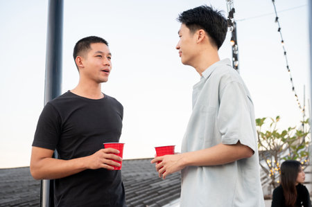Two happy young Asian male friends are enjoying talking while partying on a rooftop bar together. celebration, friendship, city life, lifestyleの写真素材