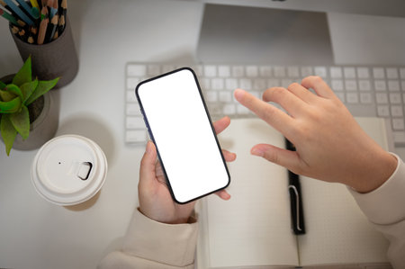 Top view image of a woman is using her smartphone at her office desk. A white-screen smartphone mockup to display your graphic ads. People and technology conceptsの写真素材