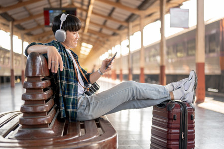 A happy and carefree Asian man backpacker is listening to music on his headphones and using his smartphone while sitting at a bench on a platform of a railway station, waiting for his train.の写真素材