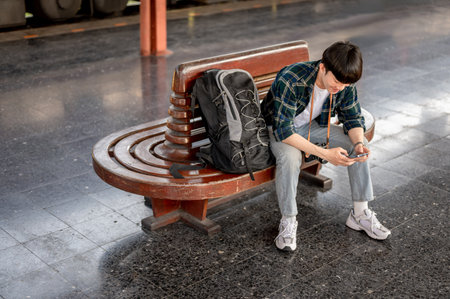 A young Asian male traveler is using his smartphone while sitting at a bench and waiting for his train at a railway station. solo traveler, backpacker, touristの写真素材