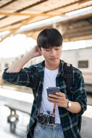 A confused young Asian tourist backpacker is checking his train ticket on his phone, checking the departure time and carriage at a railway station.の写真素材