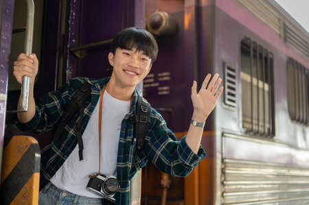 A cheerful young Asian male traveler is leaning out of the train door, smiling, waving his hand, and saying goodbye to someone while the train is leaving the railway station. vacation, solo backpackerの写真素材
