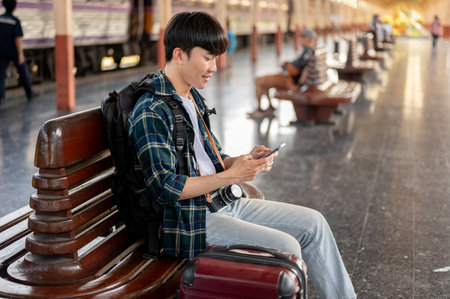 A happy young Asian man tourist is using his phone while sitting at a bench on a platform at a railway station, waiting for his train. People and commuting conceptsの写真素材