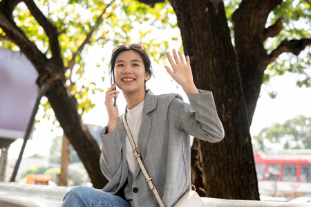 A beautiful, happy Asian woman is talking on the phone and waving her hand to her friend while she is waiting sitting on a bench in a city park. lifestyle conceptの写真素材