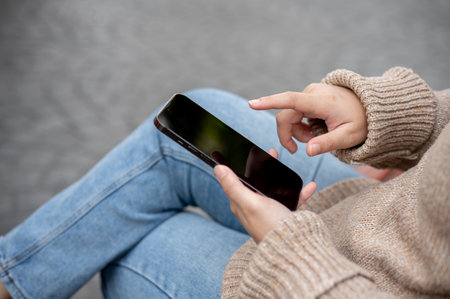 Close-up image of a woman using her smartphone while sitting at a bench outdoors in a city park, chatting, using a mobile app, or responding to messages. People and wireless technology conceptsの写真素材