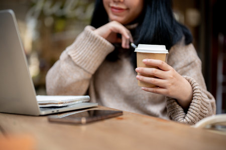 Cropped shot of a positive Asian woman holding a coffee cup, enjoying her morning coffee while working remotely at a coffee shop.の写真素材