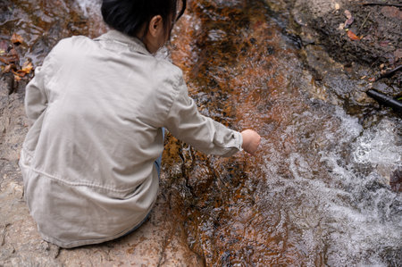 A back view image of an Asian woman sitting by the water and touching the water in a small nature canal waterfall on the mountain, feeling fresh and relaxed, washing her hands.の写真素材