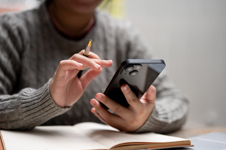 Close-up image of a woman using her smartphone while working at her desk. A female student is chatting with her friends on her phone while doing homework. People and technology conceptsの写真素材