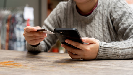 A cropped shot of a woman holding her smartphone and a credit card while sitting at a table. online...