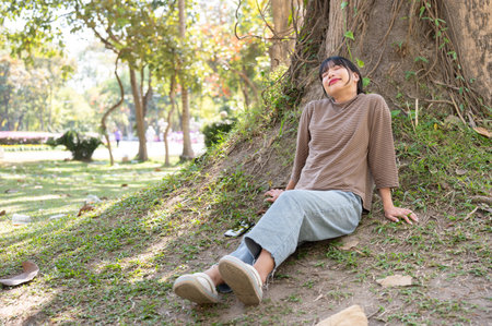 Young happy and carefree Asian woman resting under a tree in a park, eyes closed and breathing fresh air.の写真素材