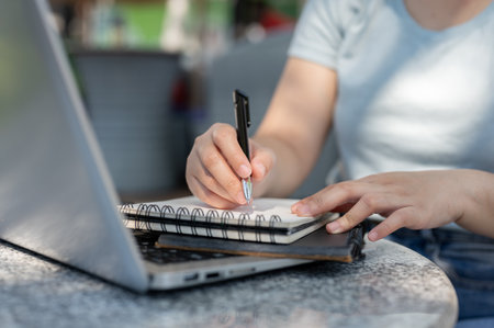 A close-up shot of a woman writing something in her notebook at an outdoor table, working remotely....