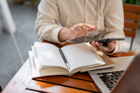 A cropped shot features a woman in a white hoodie using her smartphone while working remotely at a...