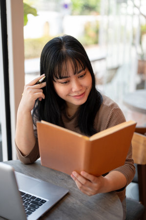A happy and positive young Asian female college student is diligently reading a book and preparing...