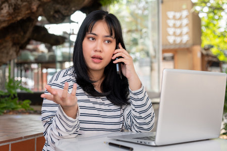 A serious Asian woman is talking on the phone with someone while working remotely at a cafe in the city. dealing business, asking for information, or explaining something on the phoneの写真素材