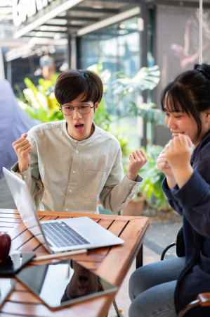 Two excited young Asian colleagues are celebrating their project success together, looking at a laptop screen with surprised faces and raising their fists.の写真素材