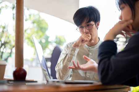 Two Asian colleagues are focusing on their project on a laptop, discussing and sharing ideas, while working remotely at a cafe in the city.の写真素材