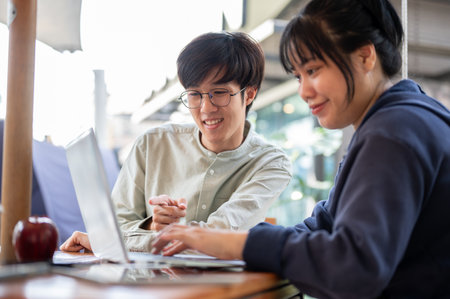 Two happy young Asian college students are discussing and working on a project in a cafe together. co-working, meeting, sharing ideasの写真素材