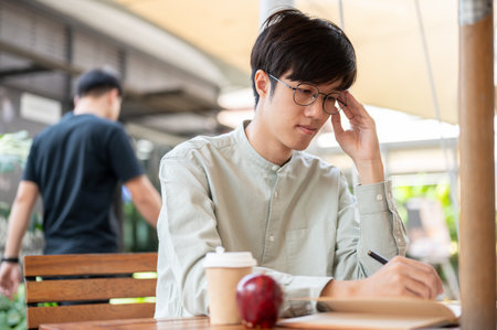 A focused and serious young Asian man is contemplating and jotting down his ideas in a book while...