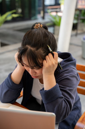 A stressed and upset young Asian woman is feeling overworked and tired while working remotely at a...