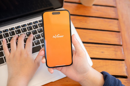 Chiang Mai, Thailand - Feb 15, 2024: A woman uses the Payoneer application on her iPhone while working remotely at a cafe. The Payoneer logo is displayed on the smartphone screen.のeditorial素材