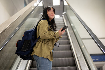A positive young Asian female solo traveler is on an escalator in the airport. departure, domestic...