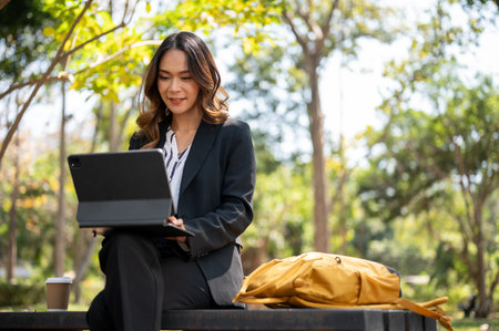 An attractive Asian businesswoman working remotely at a park, working on her digital tablet on a bench.の写真素材