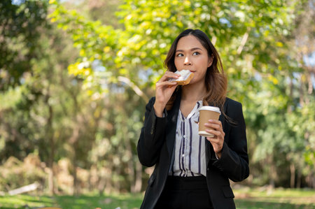 A beautiful Asian businesswoman is having a quick breakfast in a city park, eating a sandwich and drinking coffee. modern city life conceptの写真素材